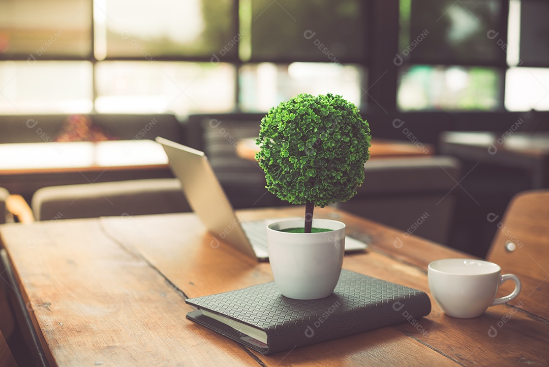 Árvore de decoração pequena, laptop, notebook e xícara de café na mesa de madeira na cafeteria.