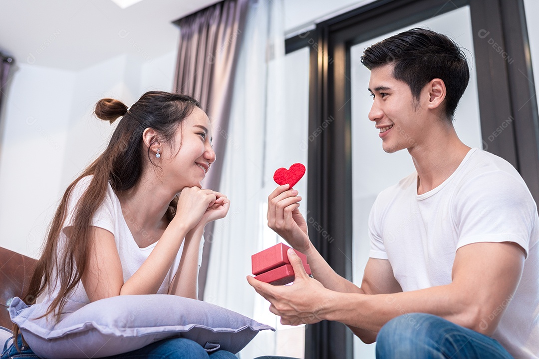 Homem segurando caixa surpresa e presente de coração vermelho para mulher no sofá em casa na sala de estar relaxe e conceito de férias.