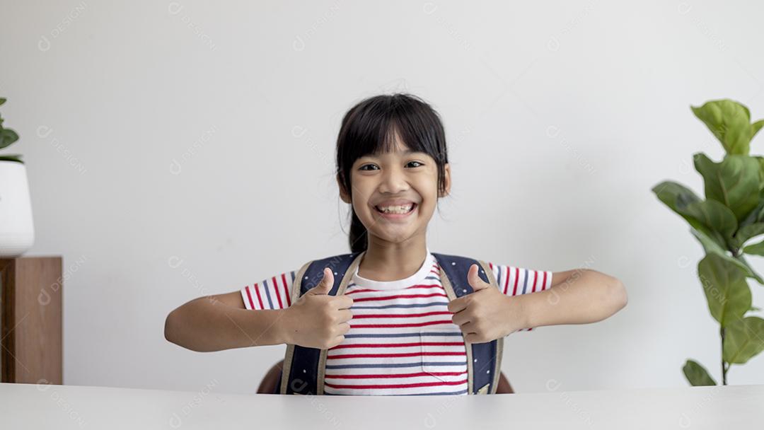 Garota asiática da escola primária com mochila mostrando os polegares.