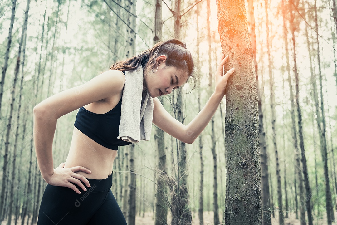 Mulher de beleza asiática cansativa de correr na floresta. Apoie-se na árvore.