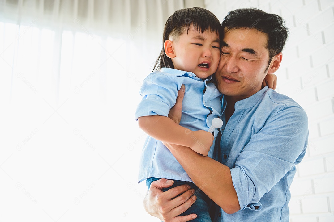 Pai japonês asiático carregando e consolando seu filho chorando no quarto em sua casa com janela e fundo de cortina branca.