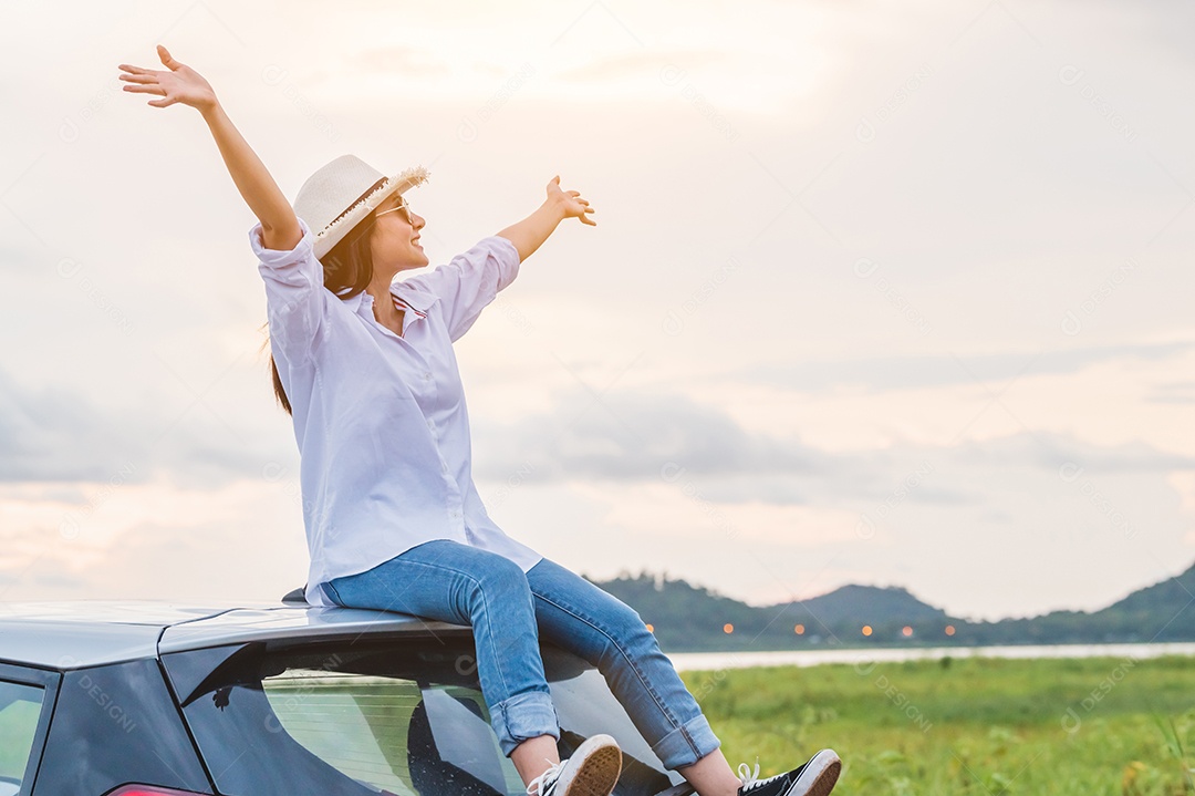 A mulher asiática feliz abriu os braços no telhado do carro sob o pôr do sol à beira-mar com o fundo da natureza durante a viagem nas férias.