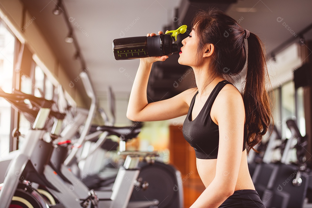 Mulher bonita asiática bebendo shake de proteína ou água potável no ginásio de treinamento esportivo.