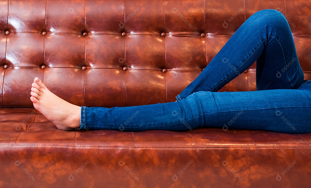 Pernas de mulher vestindo jeans enquanto relaxa no cinema ou em casa no sofá de couro marrom, relaxe e conceito de férias, conceito de dormir na hora de dormir.