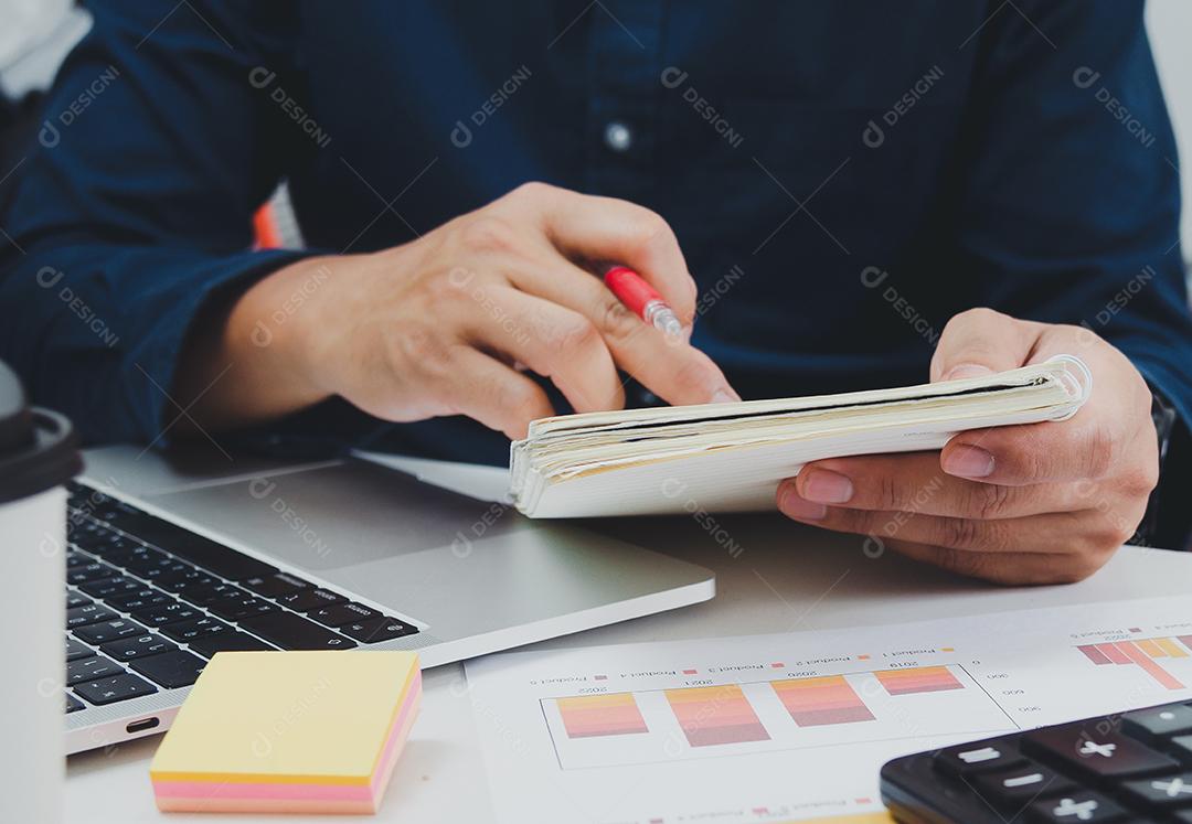 Homem de negócios, segurando a caneta e o livro na mesa, documento contábil, finanças fiscais, gráfico, relatório, investimento e marketing com computador portátil.
