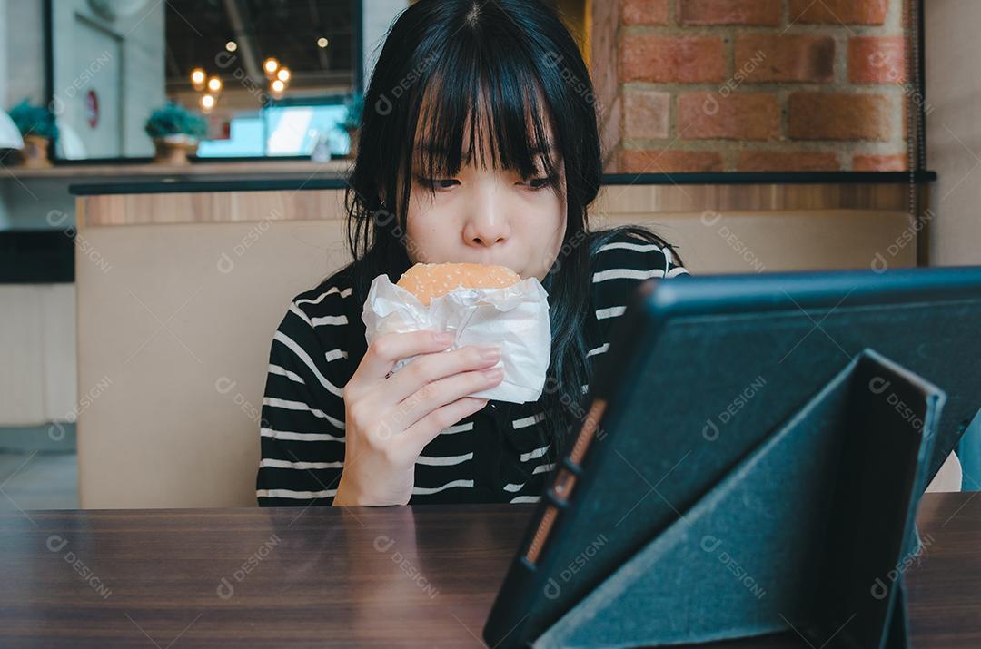 Jovem está comendo um hambúrguer e assistindo um tablet de tecnologia de mídia social online na mesa.