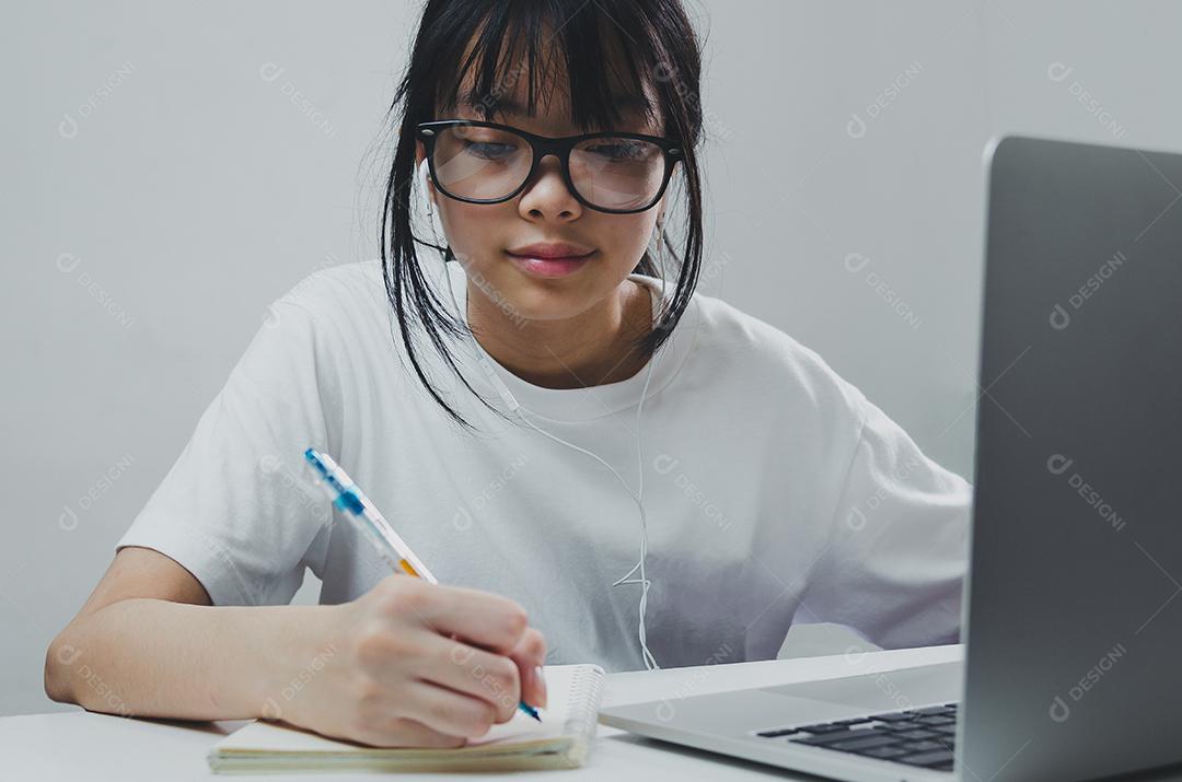 Menina segurando caneta escrevendo livro estudo aprendendo digital internet vídeo computador on-line laptop em casa.