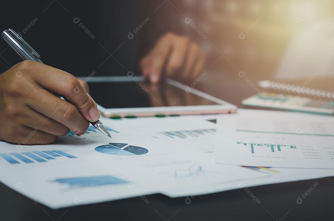 Mão de empresário segurando caneta apontando papelada gráfico relatório análise estatística e documento gráfico financeiro investimento marketing na mesa na empresa.