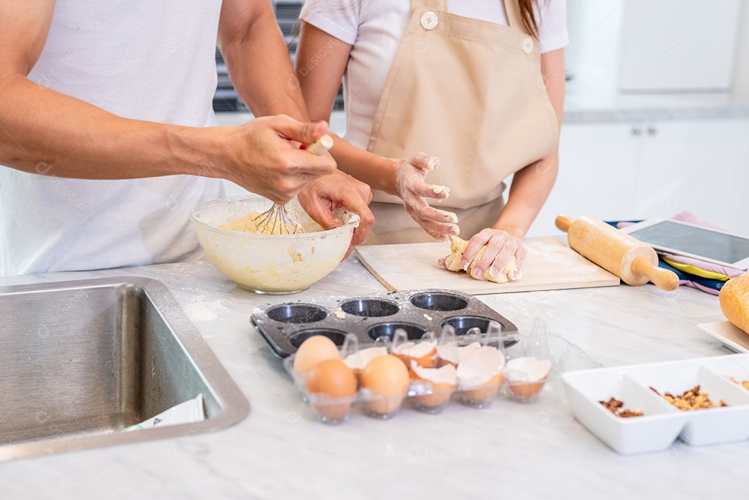 Casais asiáticos felizes cozinhando e assando bolo juntos na cozinha. Homem e mulher que procuram tablet seguem a etapa da receita em casa. Conceito de amor e felicidade. Doce lua de mel e tema do dia dos namorados