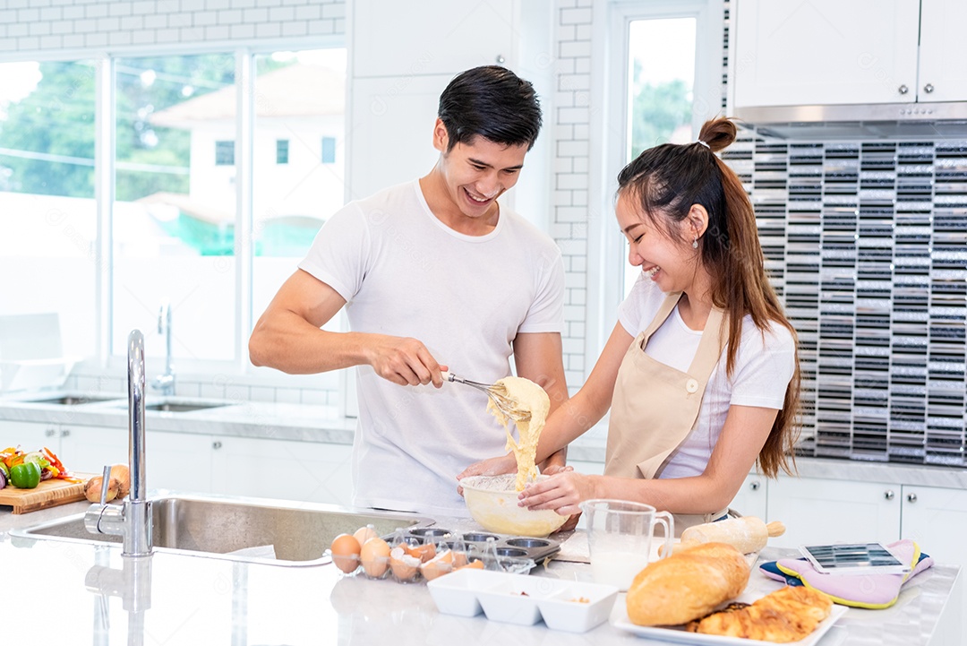 Casais asiáticos felizes cozinhando e assando bolo juntos na cozinha. Homem e mulher que procuram tablet seguem a etapa da receita em casa. Conceito de amor e felicidade. Doce lua de mel e tema do dia dos namorados.