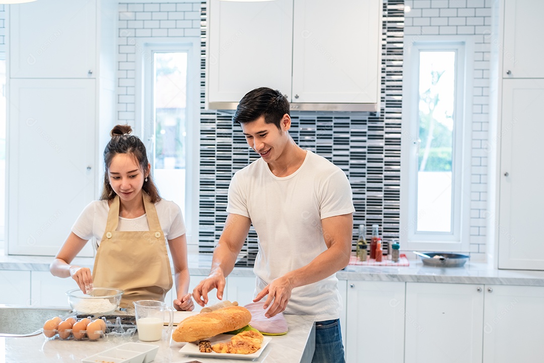 Happy Asian couples cooking and baking cake together in the kitchen. Man and woman looking for tablet follow recipe step at home. Concept of love and happiness. Sweet honeymoon and valentine's day theme.