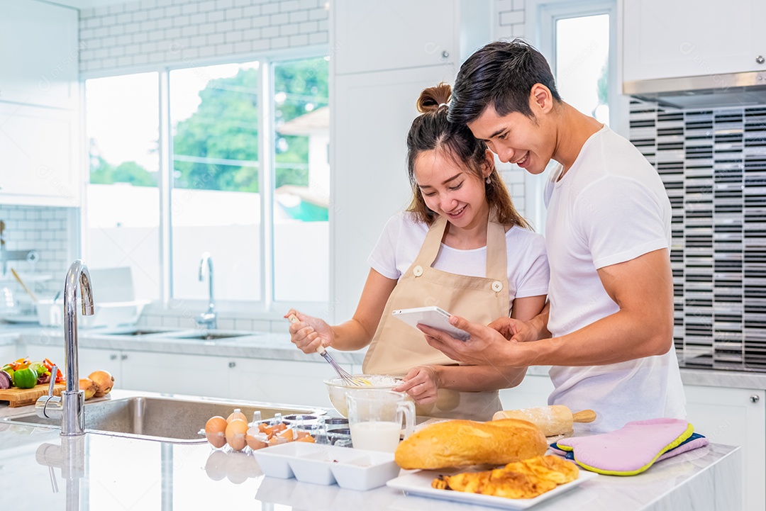Casais asiáticos felizes cozinhando e assando bolo juntos na cozinha. Homem e mulher que procuram tablet seguem a etapa da receita em casa.