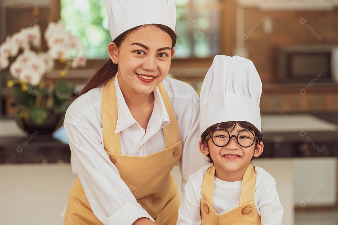 Retrato bonitinho menino asiático chef com óculos e sua mãe olhando para a câmera na cozinha caseira feliz e engraçada.