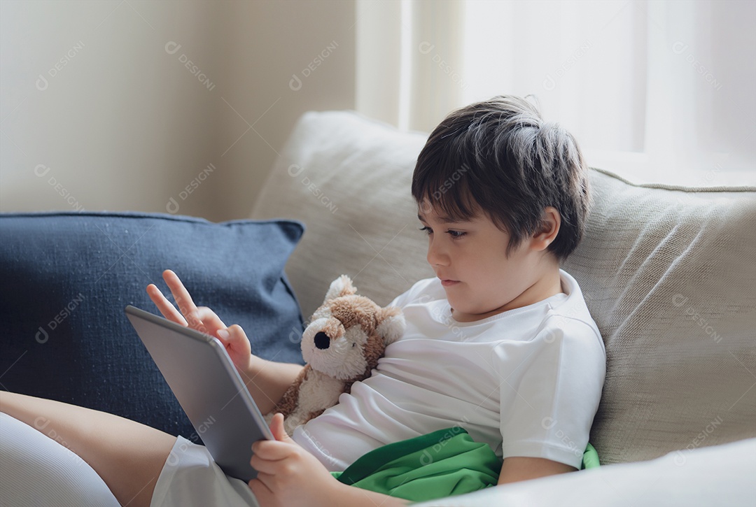 Menino usando tablet jogando jogo na internet, Criança sentada no sofá assistindo ou conversando com um amigo online
