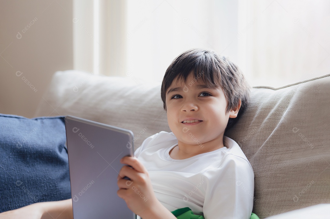 Retrato cinematográfico menino jogando no tablet sentado no sofá com a luz brilhando da janela