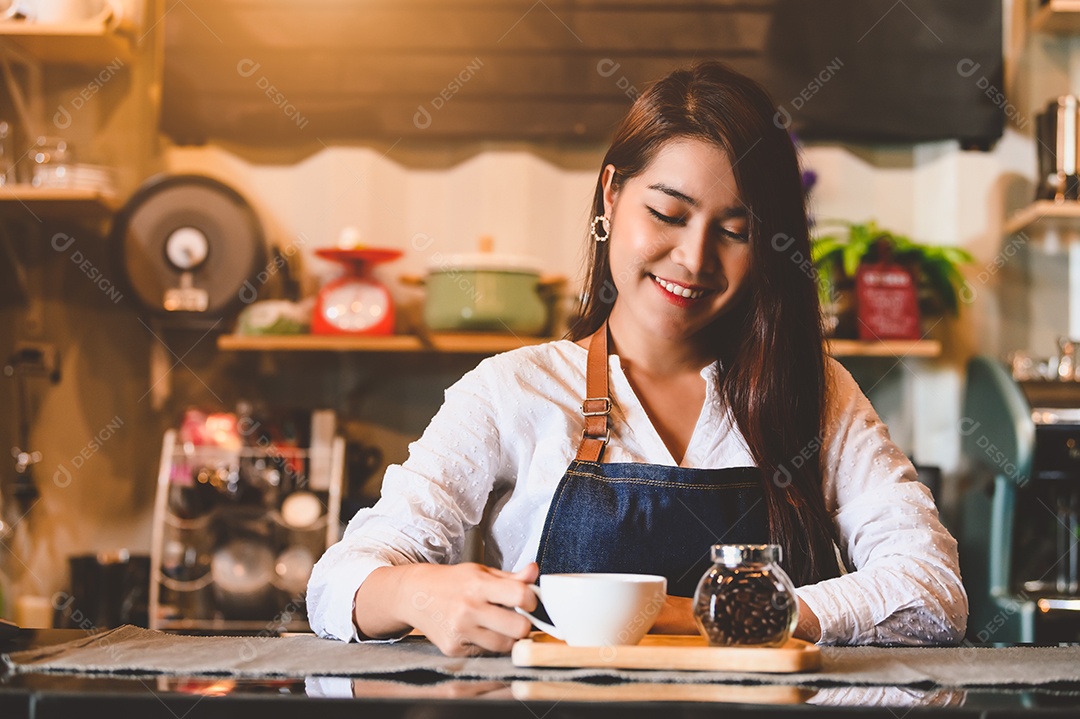 Barista feminina asiática fazendo café. Jovem segurando a xícara de café branca em pé atrás do bar do balcão do café no fundo do restaurante.
