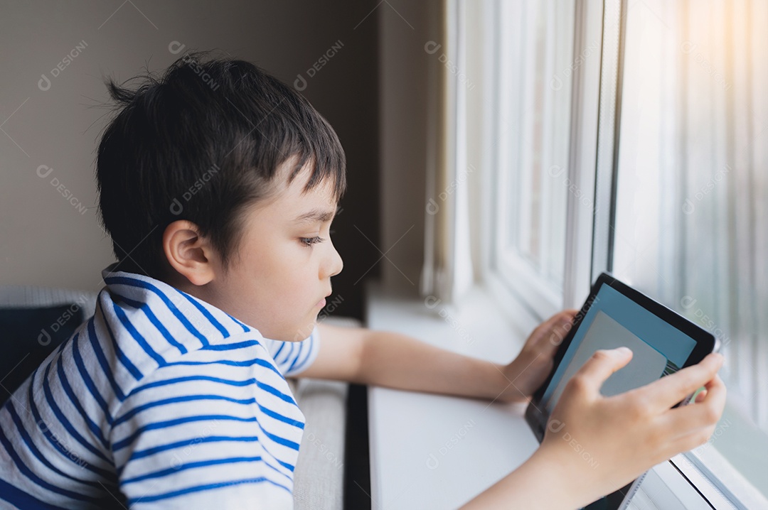 Retrato cinematográfico menino jogando jogo no tablet sentado no sofá com luz brilhando da janela