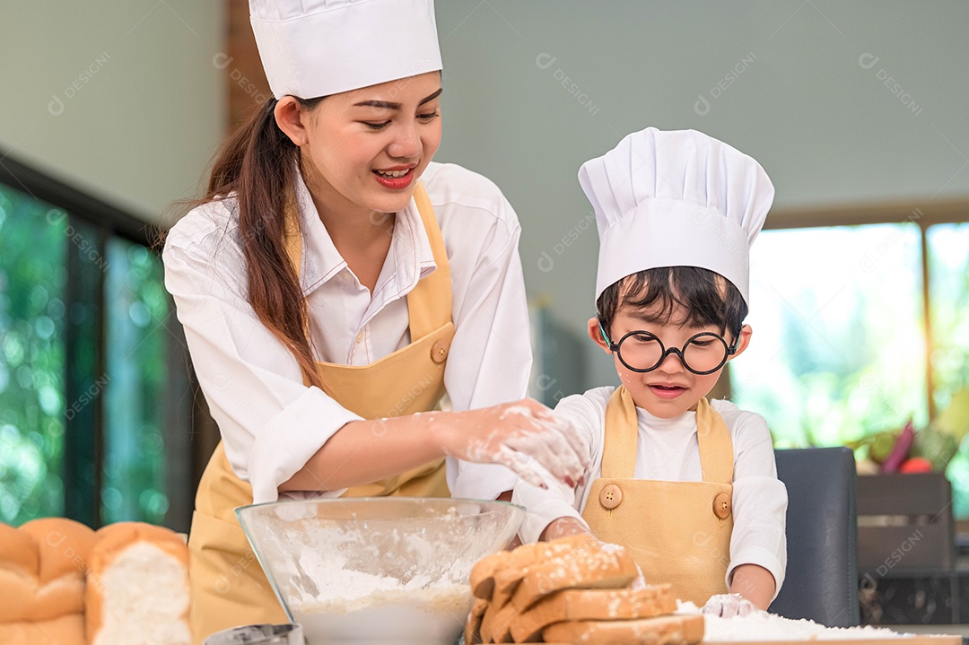 Mulher bonita e menino asiático bonitinho com óculos, chapéu de chef e avental jogando e assando padaria na cozinha de casa engraçado.