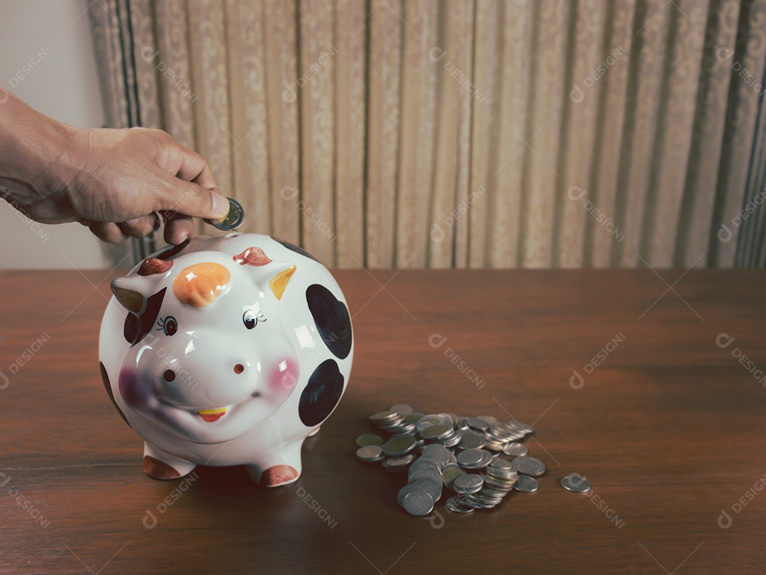 Mão colocando moedas no cofrinho de vaca, economia de dinheiro, fundo de madeira, espaço para colocar texto, conceito de economia futura