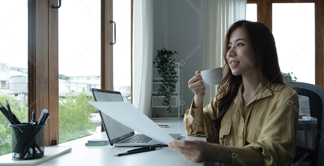 Bela jovem empresária asiática bebendo um café trabalhando no laptop no escritório.