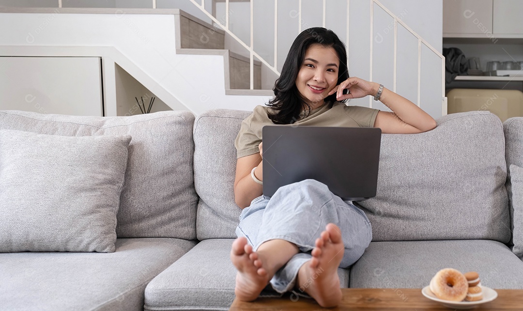 Linda mulher asiática sorridente olhando para a câmera relaxando no sofá de couro em casa.