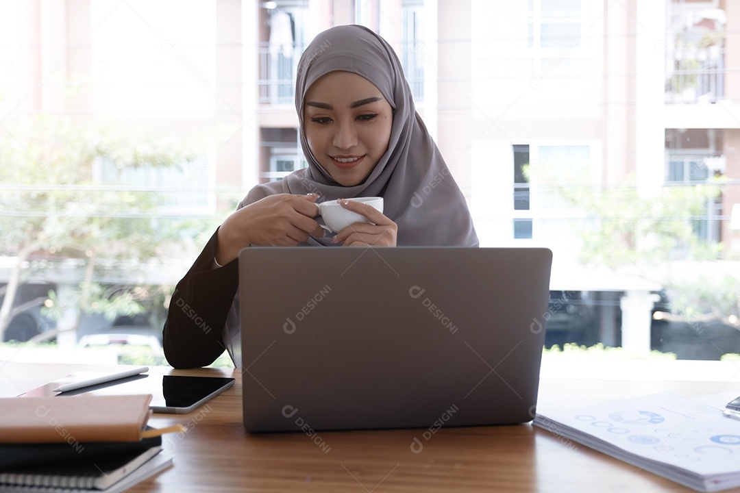 Mulher muçulmana asiática hijab segurando a xícara de café olhando para o computador