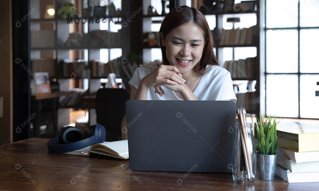 Aluna adolescente asiática sorridente estuda na biblioteca com livros e laptop fazendo pesquisas on-line para cursos