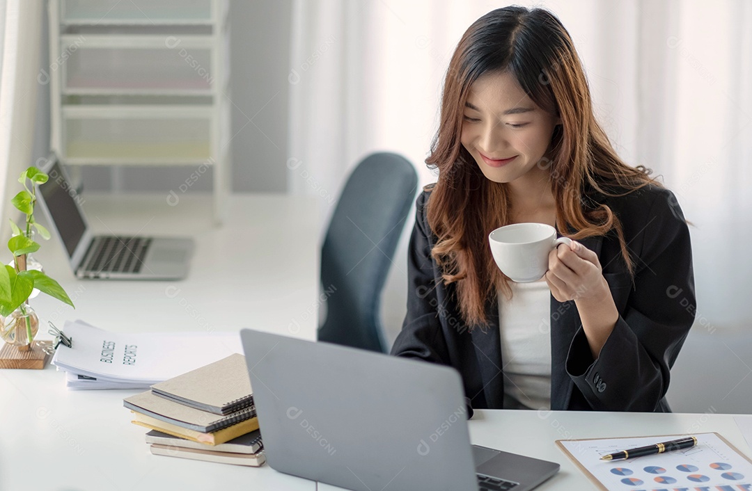 Empresária asiática sorridente segurando uma caneca de café e um laptop no escritório.