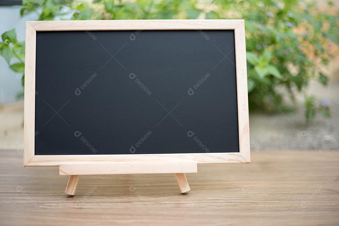 Placa preta no tampo da mesa de madeira com sol e fundo desfocado de árvore verde, modelo simulado para exibição do produto