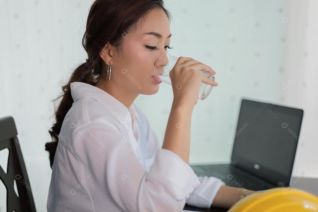 Mulher de negócios asiáticos e engenharia em vestido sentado à mesa no escritório e água potável