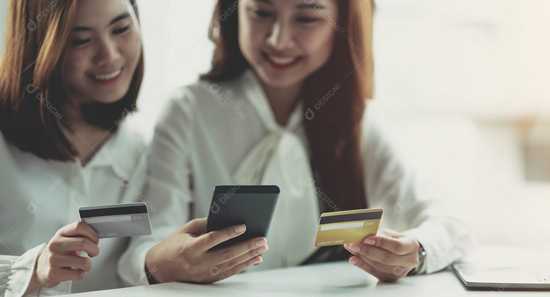 Duas jovens asiáticas seguram cartões de crédito e usam alegremente seus telefones celulares para fazer compras online.