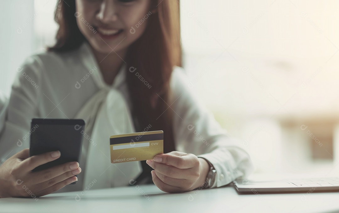 Mulher de negócios asiática feliz sentada no escritório usando um celular e segurando um cartão de crédito.
