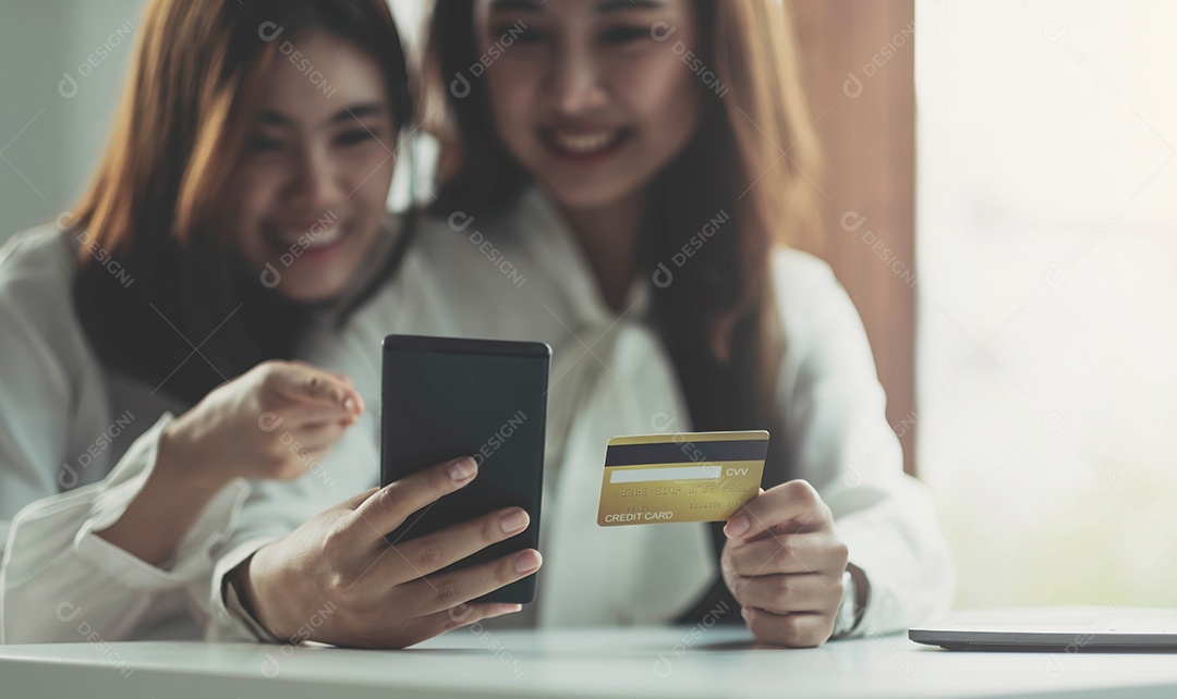 Duas jovens asiáticas seguram cartões de crédito e usam alegremente seus telefones celulares para fazer compras online.