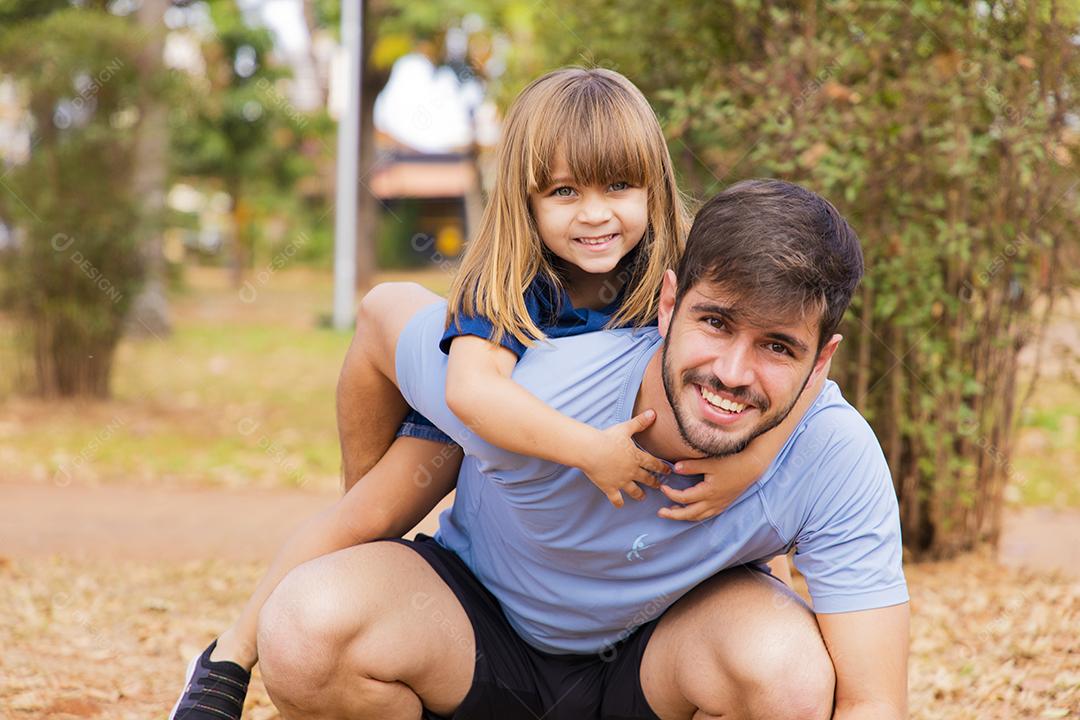 Pai e filha sorrindo para a câmera no parque. Filha sobe nas costas do papai. Feliz dia dos pais