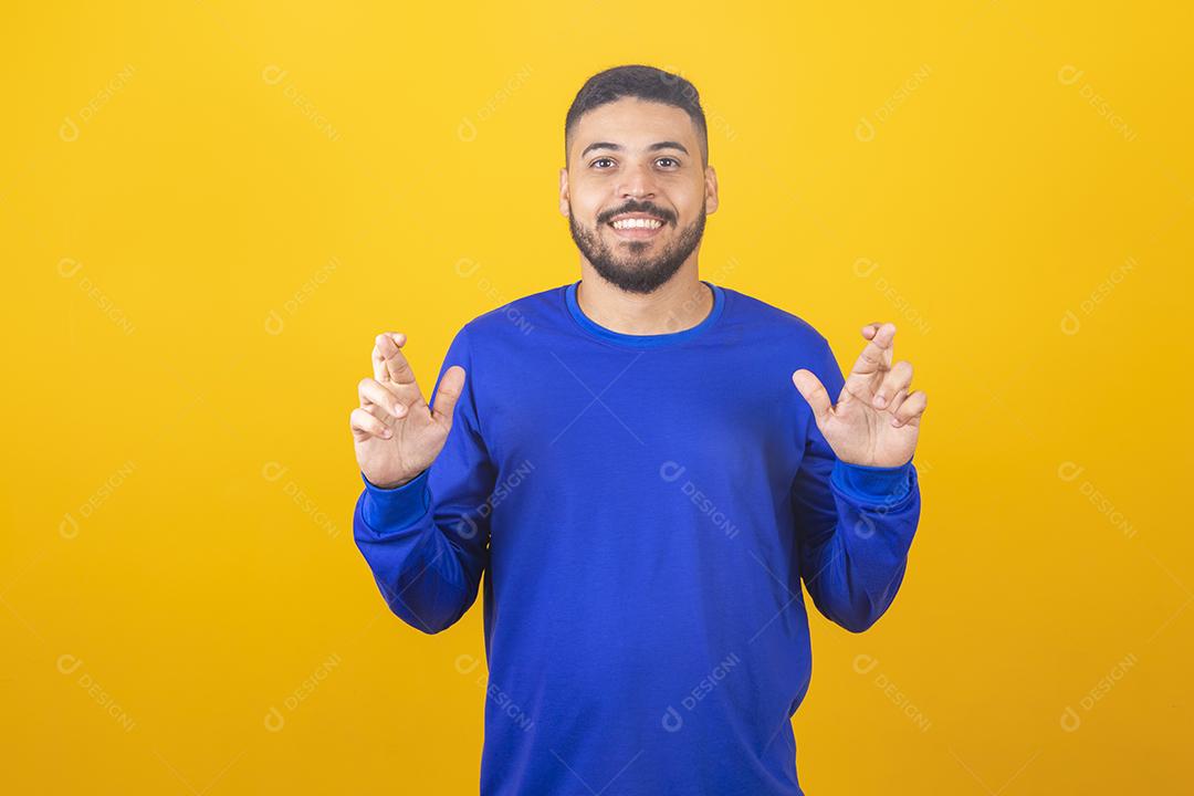 À espera de um momento especial. Retrato de jovem de camisa, mantendo os dedos cruzados sobre fundo amarelo.
