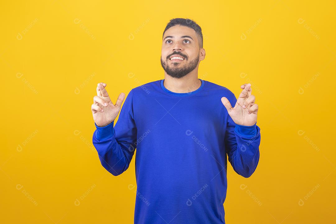 À espera de um momento especial. Retrato de jovem de camisa, mantendo os dedos cruzados sobre fundo amarelo.