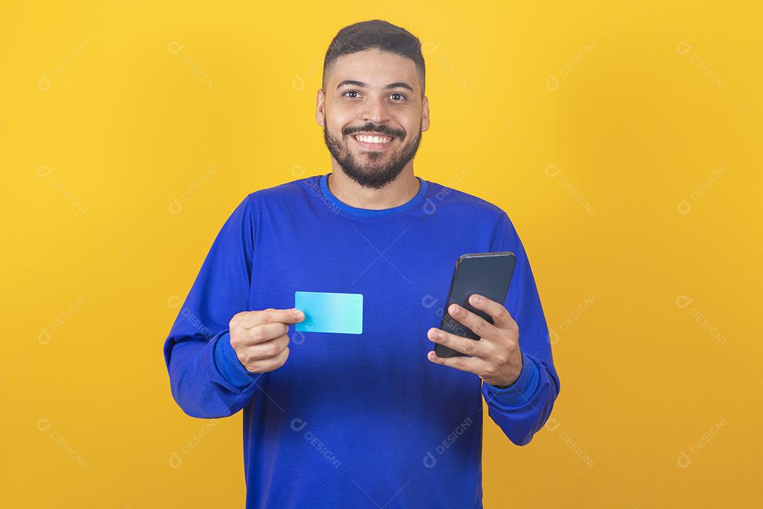 jovem bonito detém cartão de crédito e smartphone isolado em fundo amarelo. Compras online. Pagamento online.