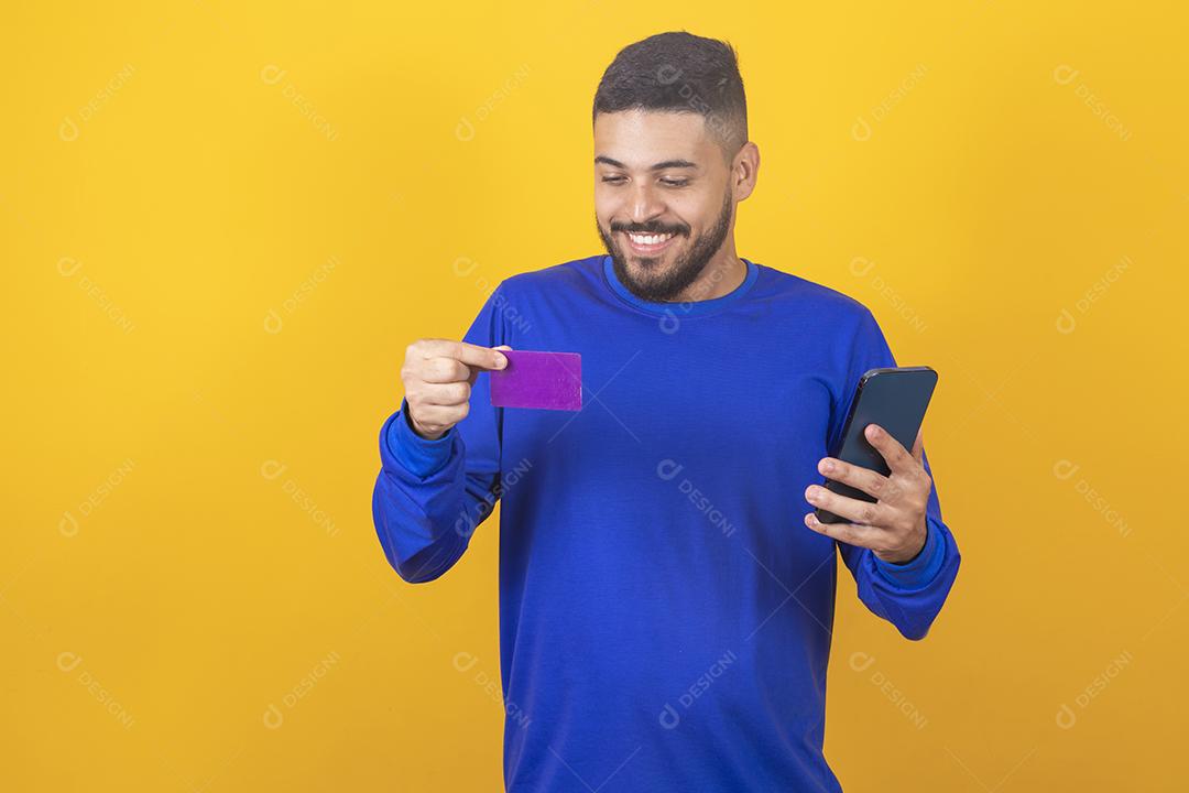jovem bonito detém cartão de crédito e smartphone isolado em fundo amarelo. Compras online. Pagamento online.