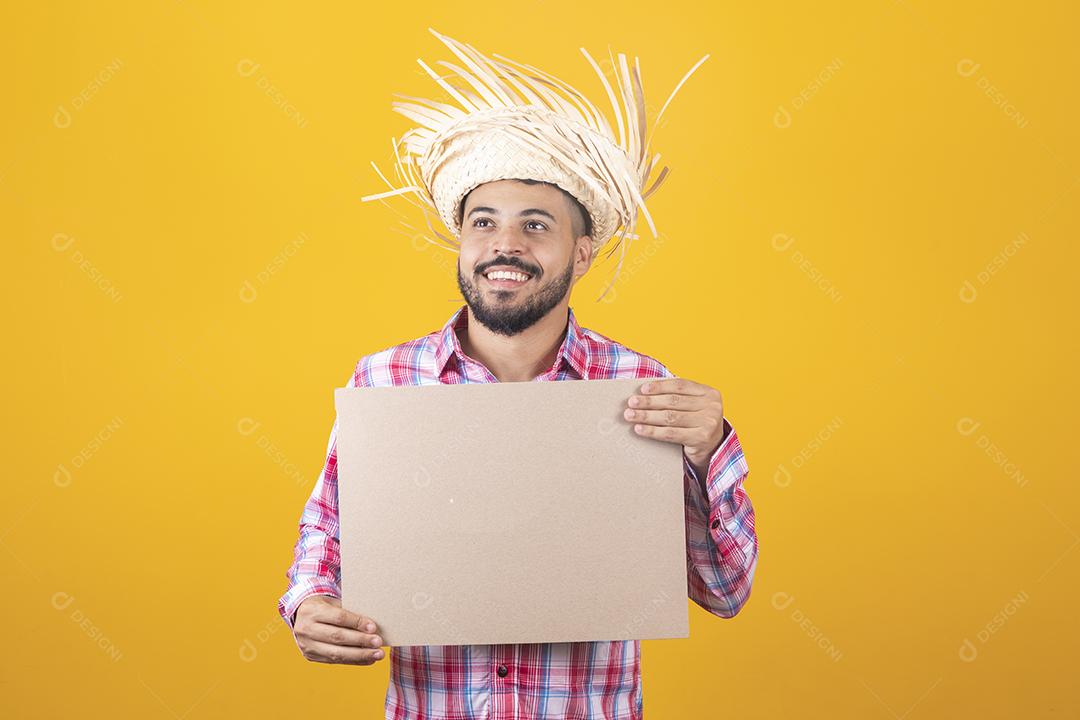 Homem brasileiro vestindo roupas típicas para a Festa Junina segurando placa
