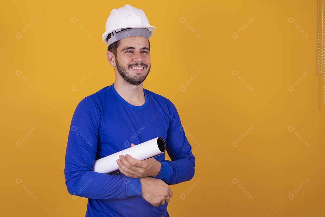 Homem jovem arquiteto com capacete e segurando plantas isoladas em fundo amarelo, apontando o dedo para o lado