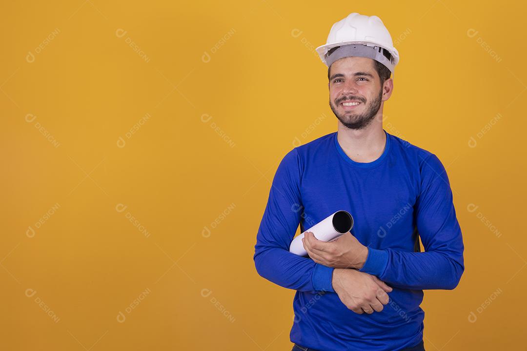 Homem jovem arquiteto com capacete e segurando plantas isoladas em fundo amarelo, apontando o dedo para o lado