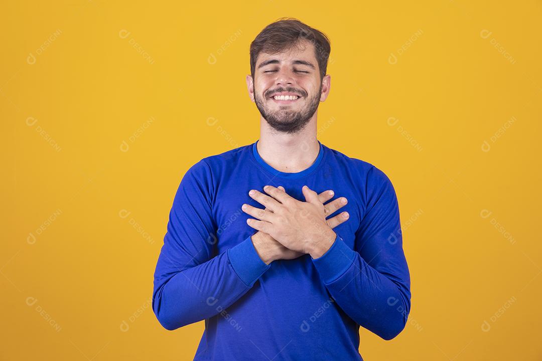 Jovem rapaz bonito tem as mãos no peito perto do coração. Emoções humanas, sentimentos reais e conceito de expressão facial.