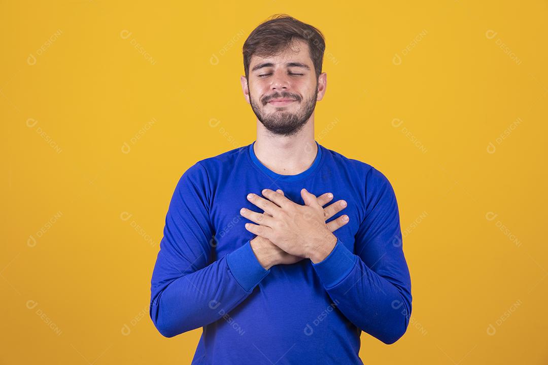 Jovem rapaz bonito tem as mãos no peito perto do coração. Emoções humanas, sentimentos reais e conceito de expressão facial.