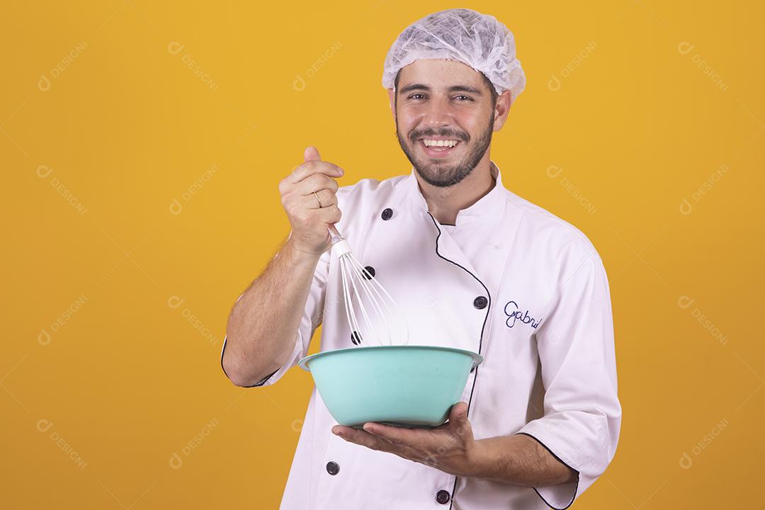 Sorrindo jovem chef masculino cozinheiro ou padeiro na camisa uniforme branca posando isolado no retrato de estúdio de fundo amarelo. Cozinhar o conceito de comida. Mock up espaço de cópia. Chicotear ovos batendo na tigela