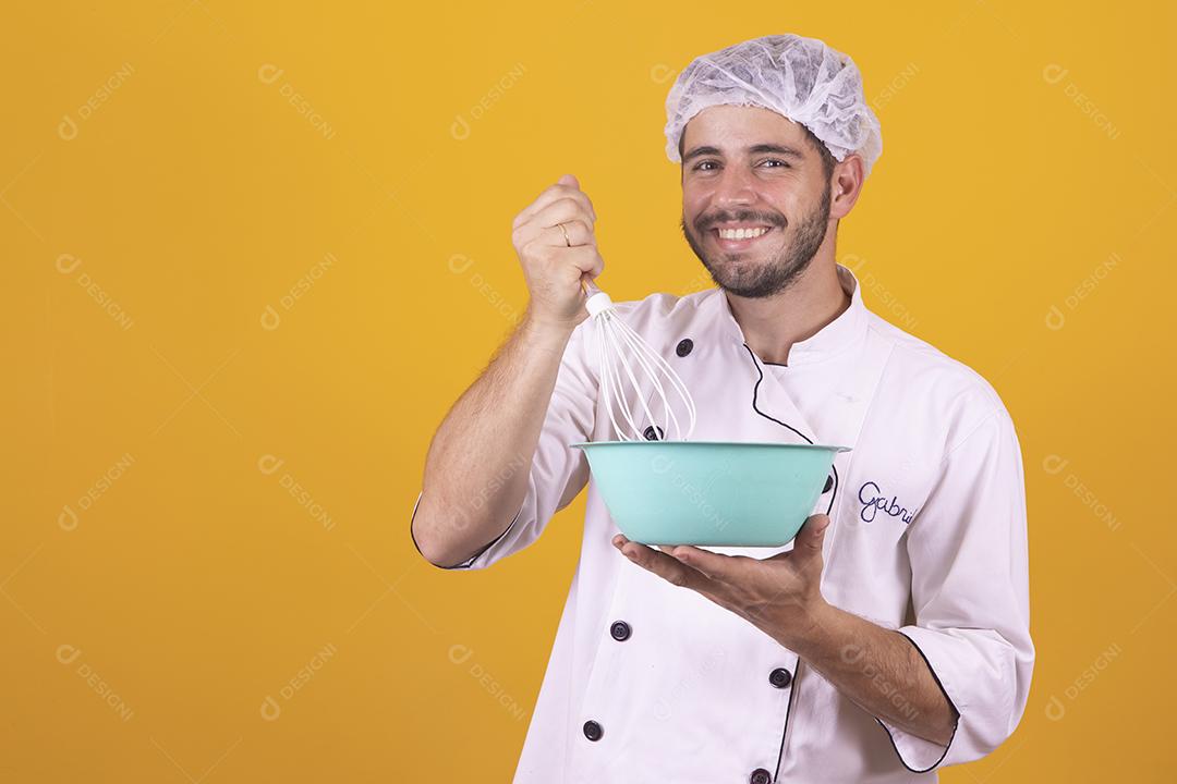 Sorrindo jovem chef masculino cozinheiro ou padeiro na camisa uniforme branca posando isolado no retrato de estúdio de fundo amarelo. Cozinhar o conceito de comida Chicotear ovos batendo na tigela