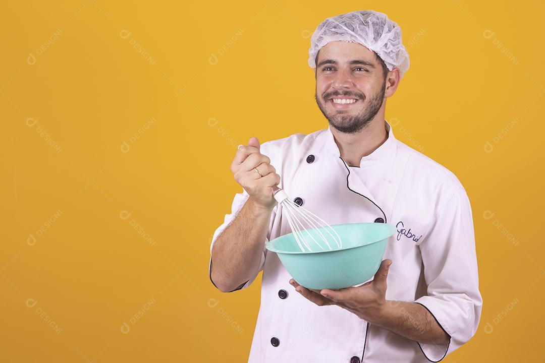 Sorrindo jovem chef masculino cozinheiro ou padeiro na camisa uniforme branca posando isolado no retrato de estúdio de fundo amarelo. Cozinhar o conceito de comida. Mock up espaço de cópia. Chicotear ovos batendo na tigela