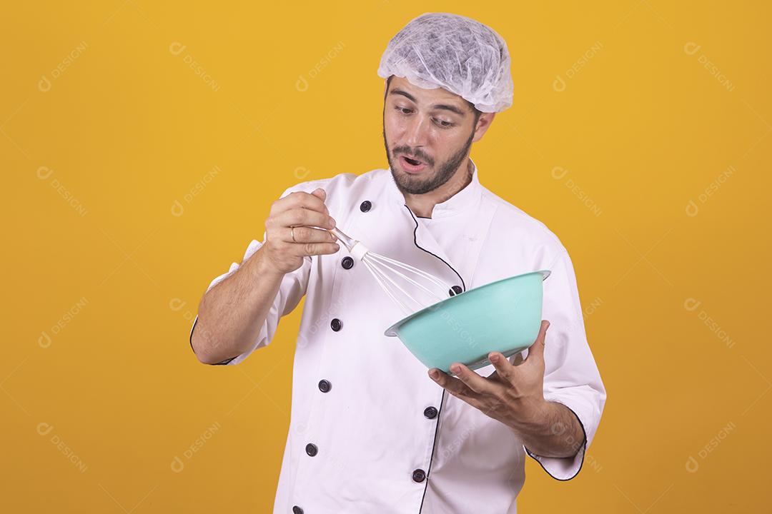 Sorrindo jovem chef masculino cozinheiro ou padeiro na camisa uniforme branca posando isolado no retrato de estúdio de fundo amarelo. Cozinhar o conceito de comida. Mock up espaço de cópia. Chicotear ovos batendo na tigela