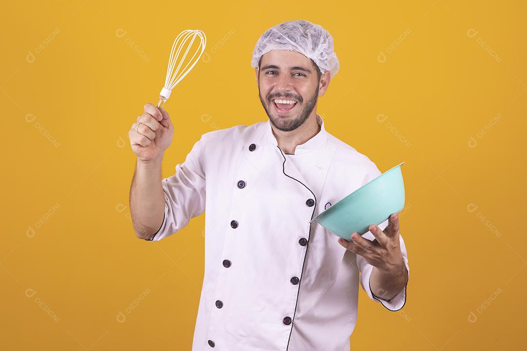 Sorrindo jovem chef masculino cozinheiro ou padeiro na camisa uniforme branca posando isolado no retrato de estúdio de fundo amarelo. Cozinhar o conceito de comida. Mock up espaço de cópia. Chicotear ovos batendo na tigela