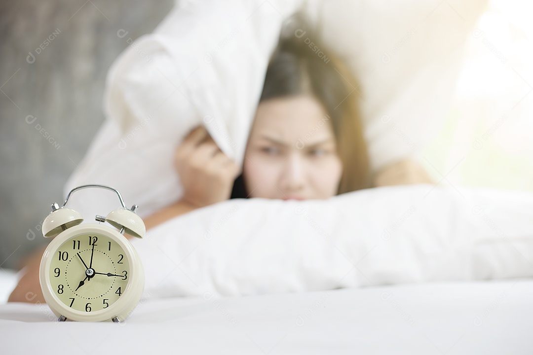 Mulher asiática odeia acordar cedo de manhã. Menina com sono olhando para o despertador e tentando se esconder debaixo do travesseiro