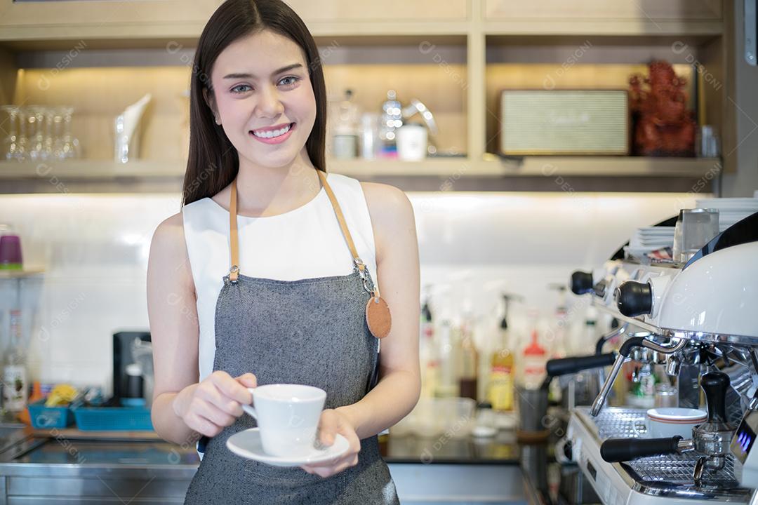 Mulher asiática Barista sorrindo e segurando a xícara de café e usando a máquina no balcão da cafeteria